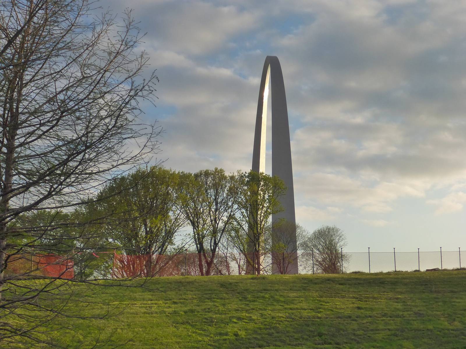 The Gateway Arch at sunrise seen across the grounds, with spring trees and a grass slope in the foreground.